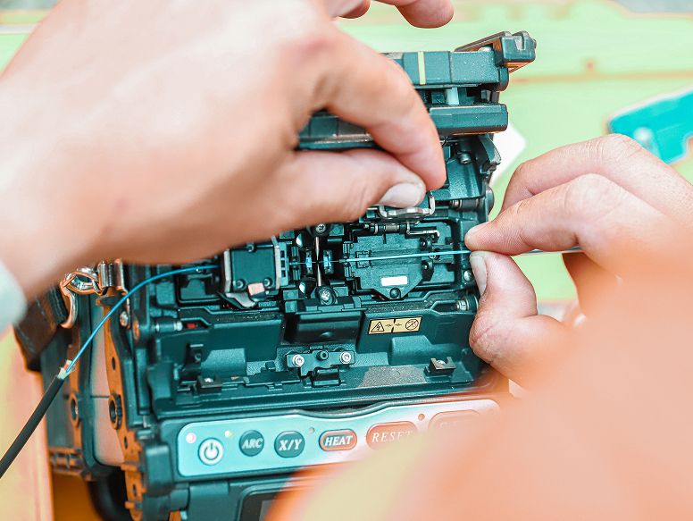 Nahaufnahme von Händen, die ein Gerät reparieren, wobei der Schwerpunkt auf komplizierten Komponenten und Verdrahtungen in einem elektronischen Gerät auf einer hellen Oberfläche liegt.