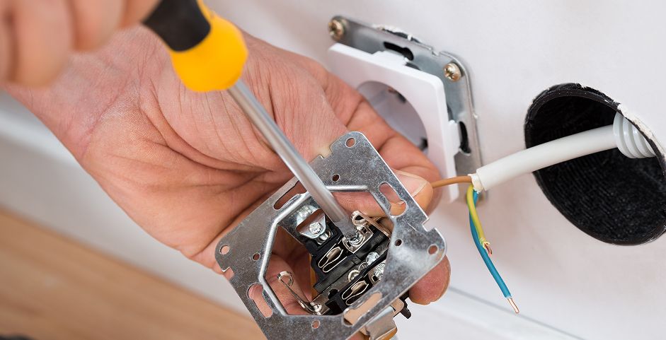 Eine Person benutzt einen Schraubenzieher, um eine Steckdose in einer Wand zu installieren, wobei die Kabel hinter der Steckdose sichtbar sind.